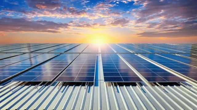 VÁRIAS PLACAS DE ENERGIA SOLAR FOTOVOLTAICA SOB O TELHADO DE UMA GRANDE EMPRESA E AO FUNDO UM PÔR DO SOL