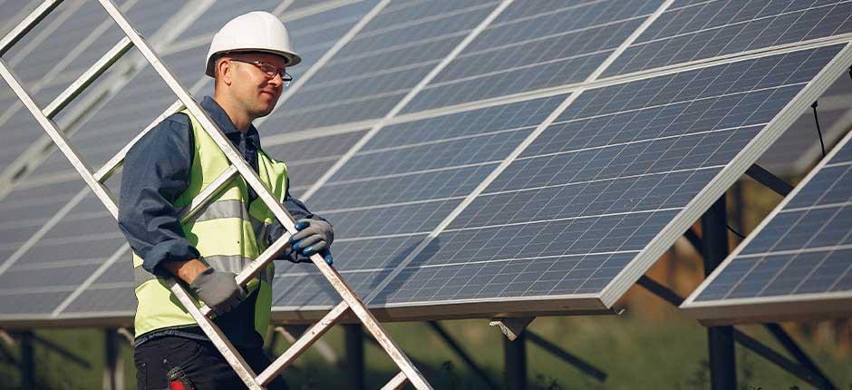 TRABALHADOR DE ENERGIA SOLAR AO AR LIVRE E SEGURANDO UMA ESCADA, EM FRENTE A UMA SÉRIE DE PAINÉIS SOLARES, ELE ESTÁ USANDO UM CAPACETE BRANCO, UM COLETE DE SEGURANÇA REFLETIVO E LUVAS DE PROTEÇÃO